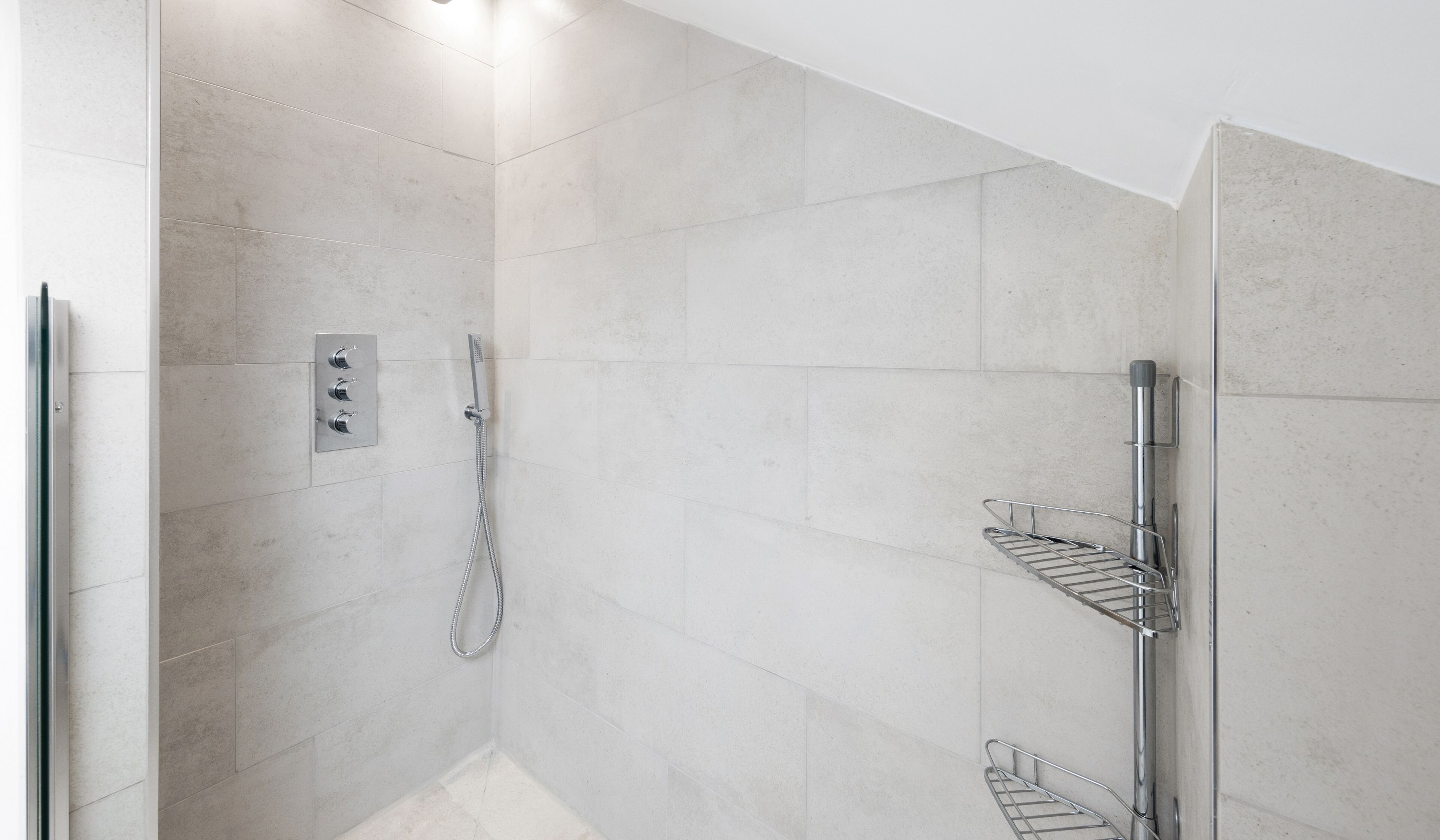 Un área de ducha moderna con ducha de mano, panel de control en la pared y repisa metálica.