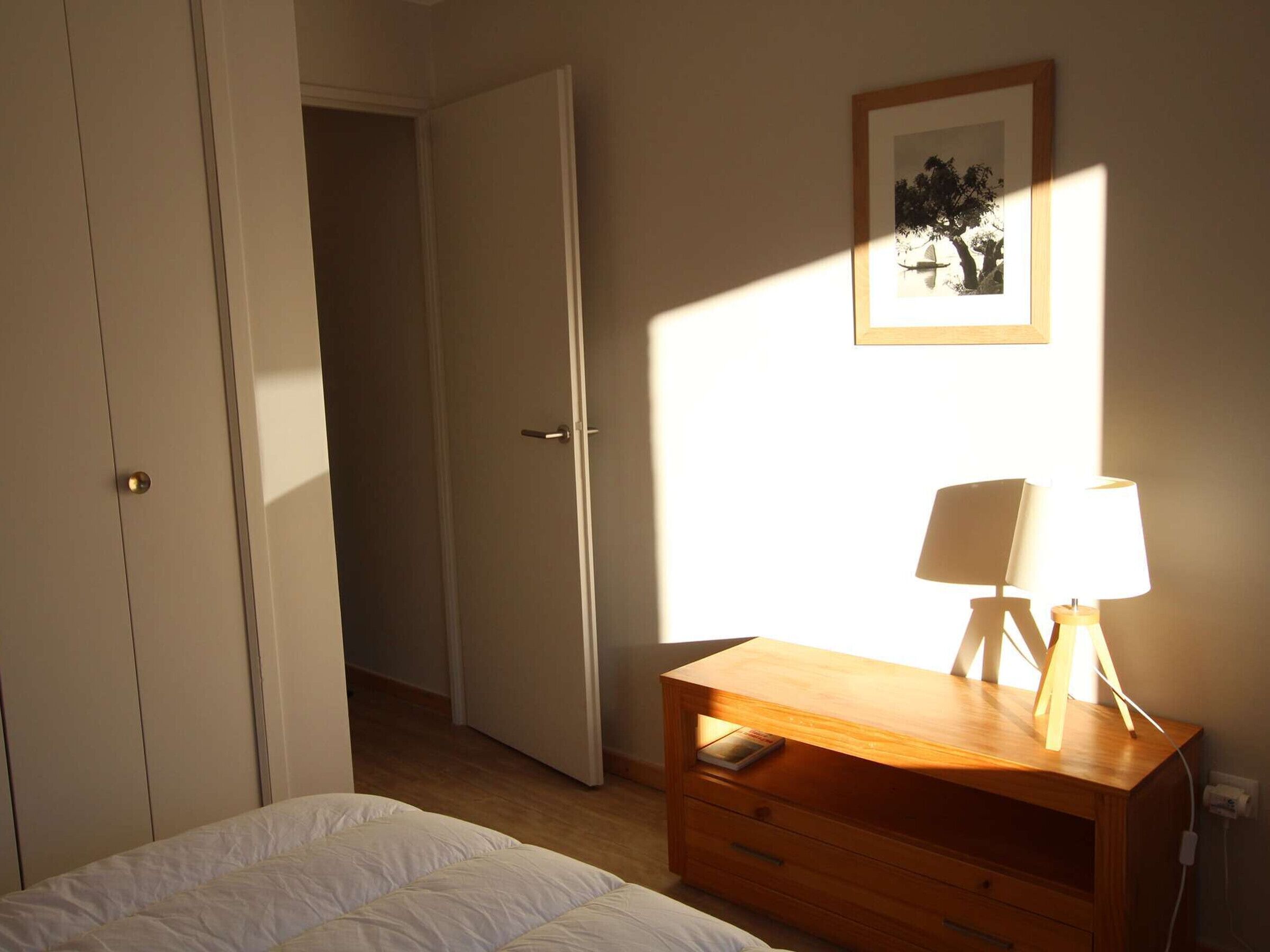 A bedroom with a bed, a wooden dresser with a lamp, a door, and a framed picture on the wall.