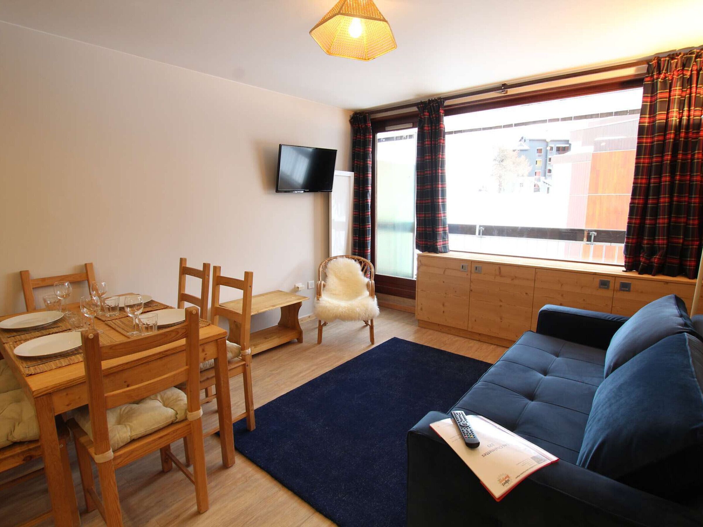 A living room with a sofa, a dining table with chairs, a television mounted on the wall, and a window with curtains.