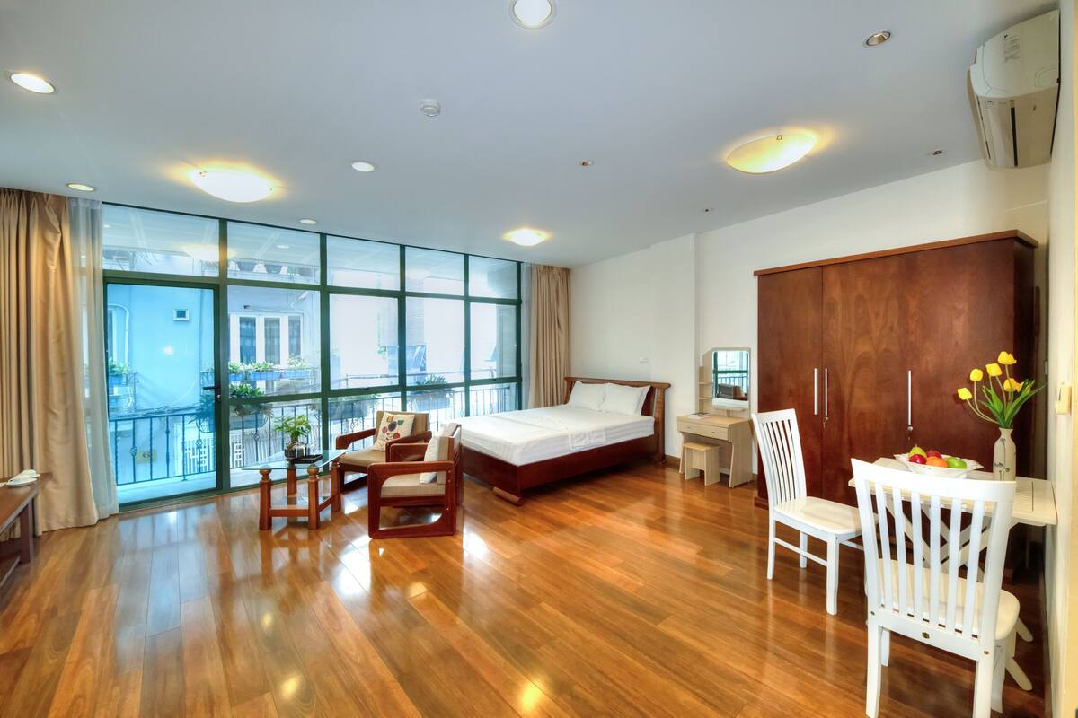 A hotel room with a large bed, a desk, a chair, and a wardrobe. There is a window with a view of buildings outside.