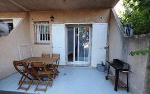 A covered patio with a wooden table and chairs, a sliding glass door, and a small outdoor grill. - Welcome to Filippi Furnished Bungalows! (PORTO-VECCHIO)