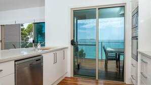 Une cuisine moderne équipée d’un lave-vaisselle, de meubles blancs et d’une porte-fenêtre coulissante donnant sur un balcon avec vue sur la mer.
