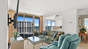 Un balcon avec vue sur l’océan et les bâtiments, un canapé, une chaise, une table et un petit coin cuisine.