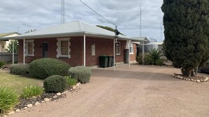 A single-story brick building with a covered porch, a gravel driveway, and a large coniferous tree. - Paradise @ Port Hughes, 500M walk to beach, ALL LINEN & TOWELS PROVIDED (Port Hughes)