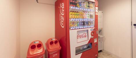 A Coca-Cola vending machine with a selection of beverages, two red trash bins, and a white refrigerator in a room with beige walls and a tiled floor.