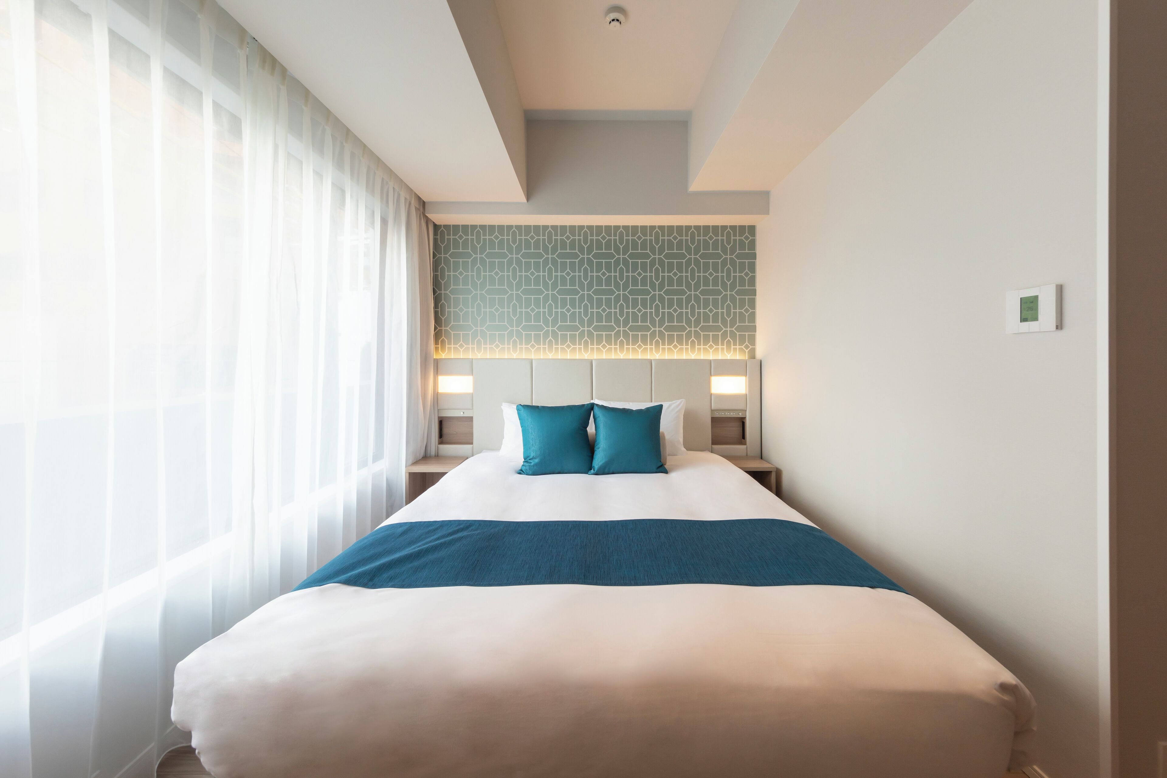 A hotel room with a large bed, a headboard with a geometric pattern, and a bedside table with a lamp.