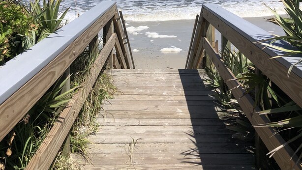 Ein hölzerner Steg führt zu einem Strand, wo Wellen an das Ufer brechen.