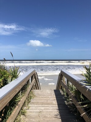 Ein hölzerner Steg führt zu einem Strand, wo Wellen an das Ufer brechen.