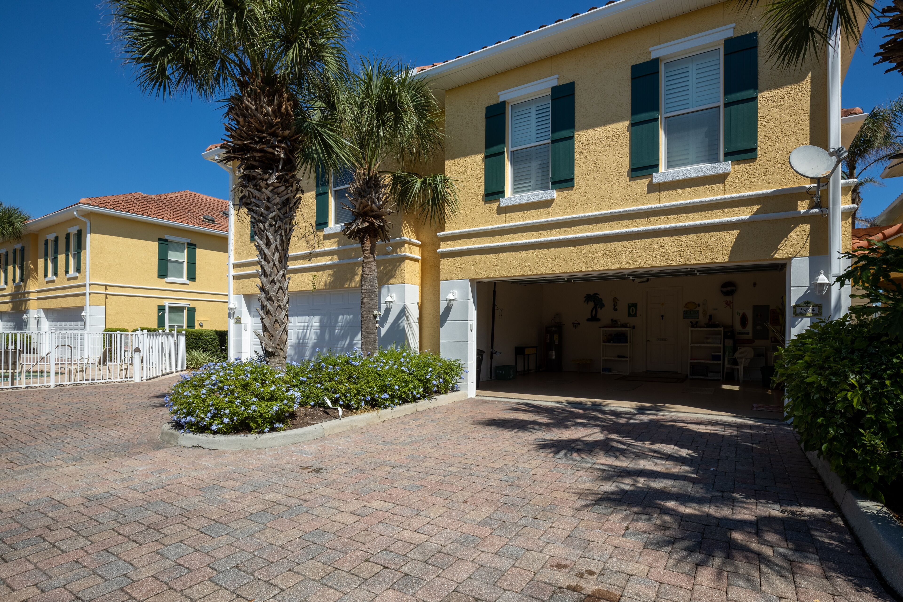 A yellow building with green shutters, a paved driveway, and a palm tree.