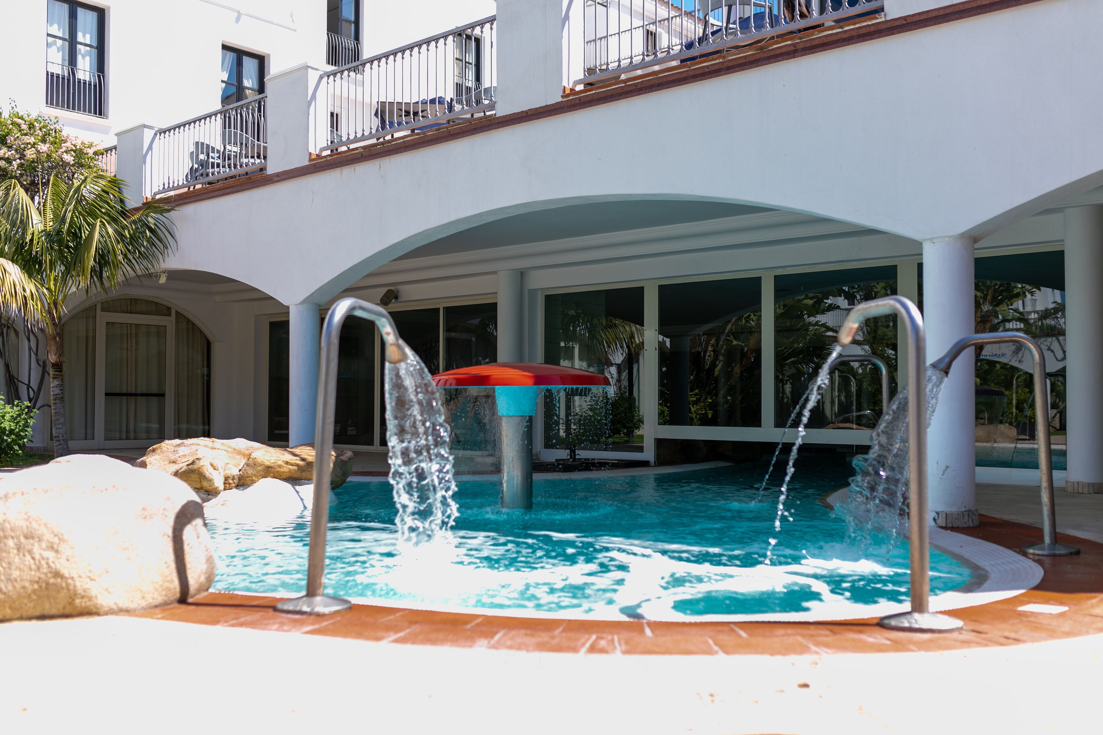 A pool with two water jets, a red umbrella, and a building with balconies and large windows.