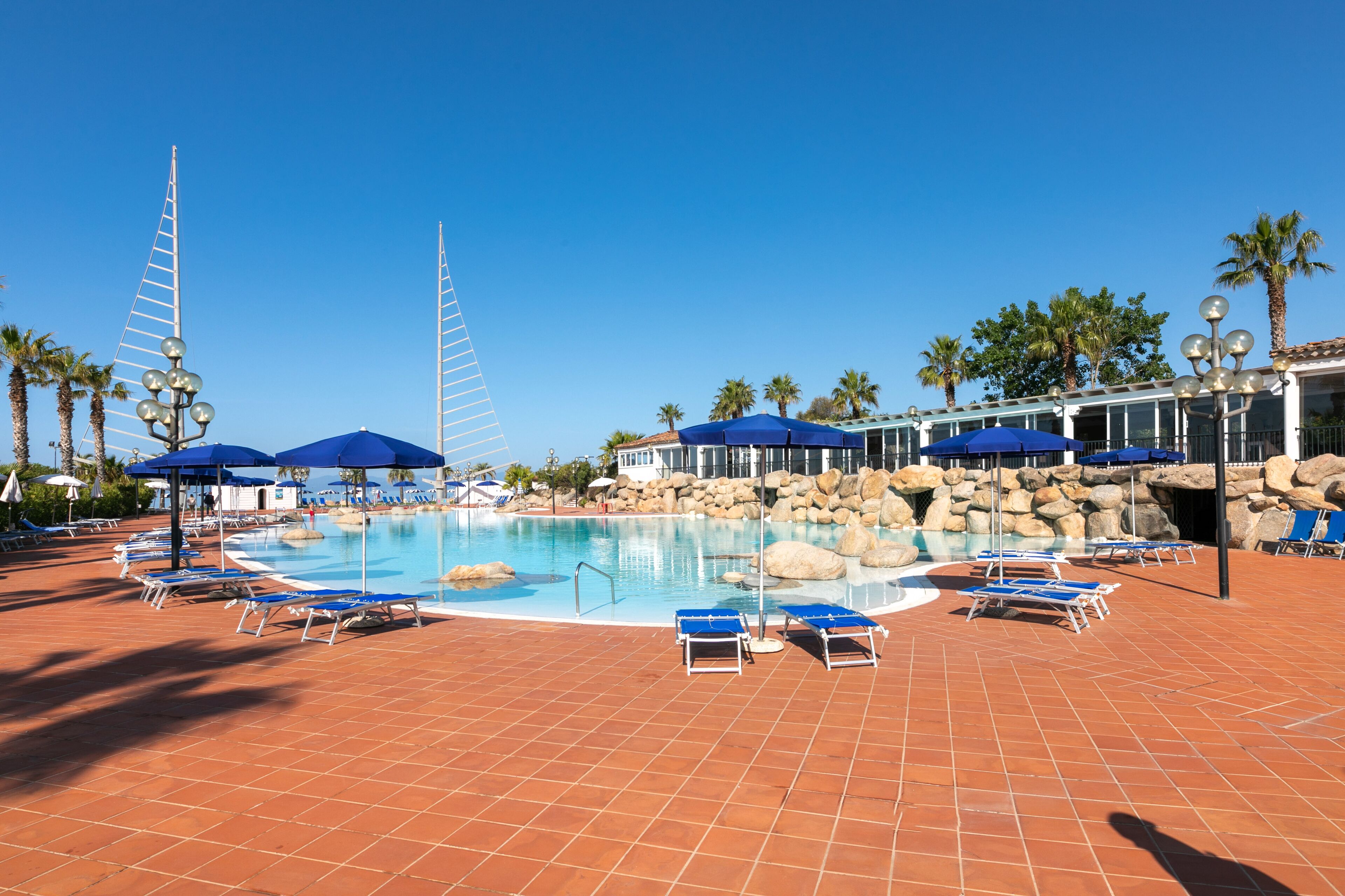 Zona de piscina con sombrillas azules, sillas de descanso y un cielo azul despejado.