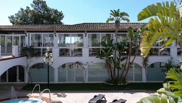 Uma área de piscina com espreguiçadeiras, uma fonte de água e um edifício com janelas amplas e portas em arco.