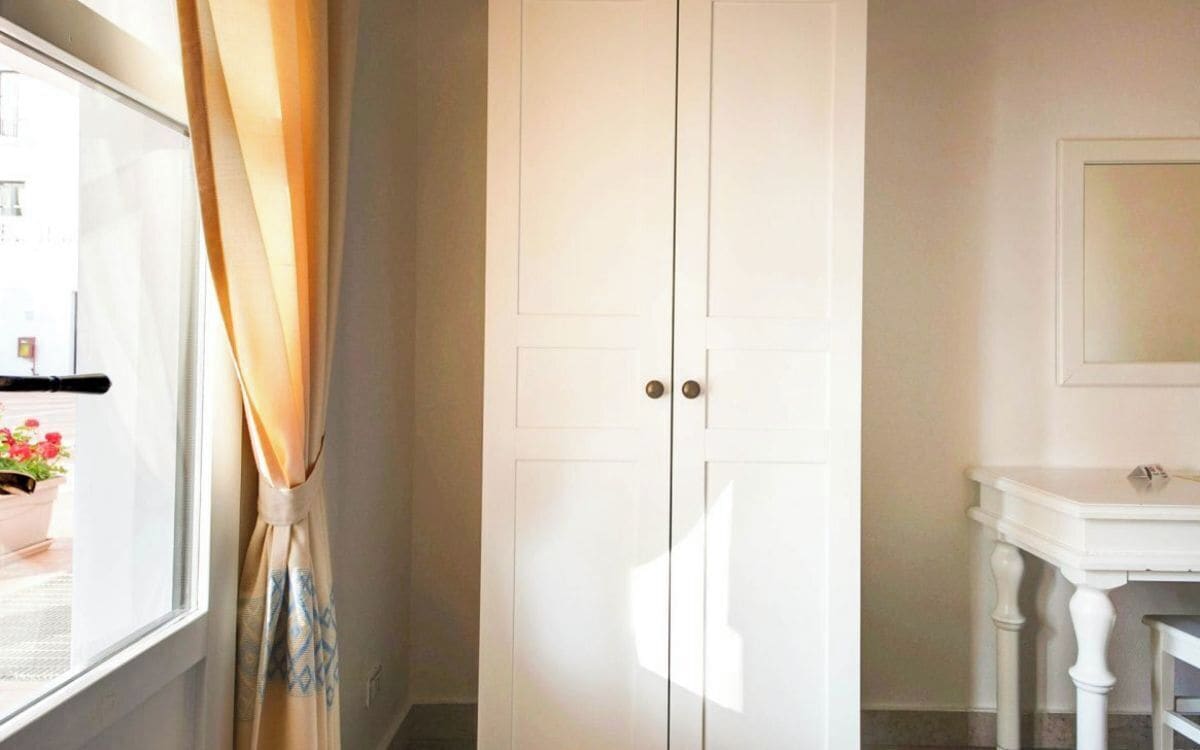 A white wardrobe with two doors, a white vanity table with a mirror, and a window with curtains.