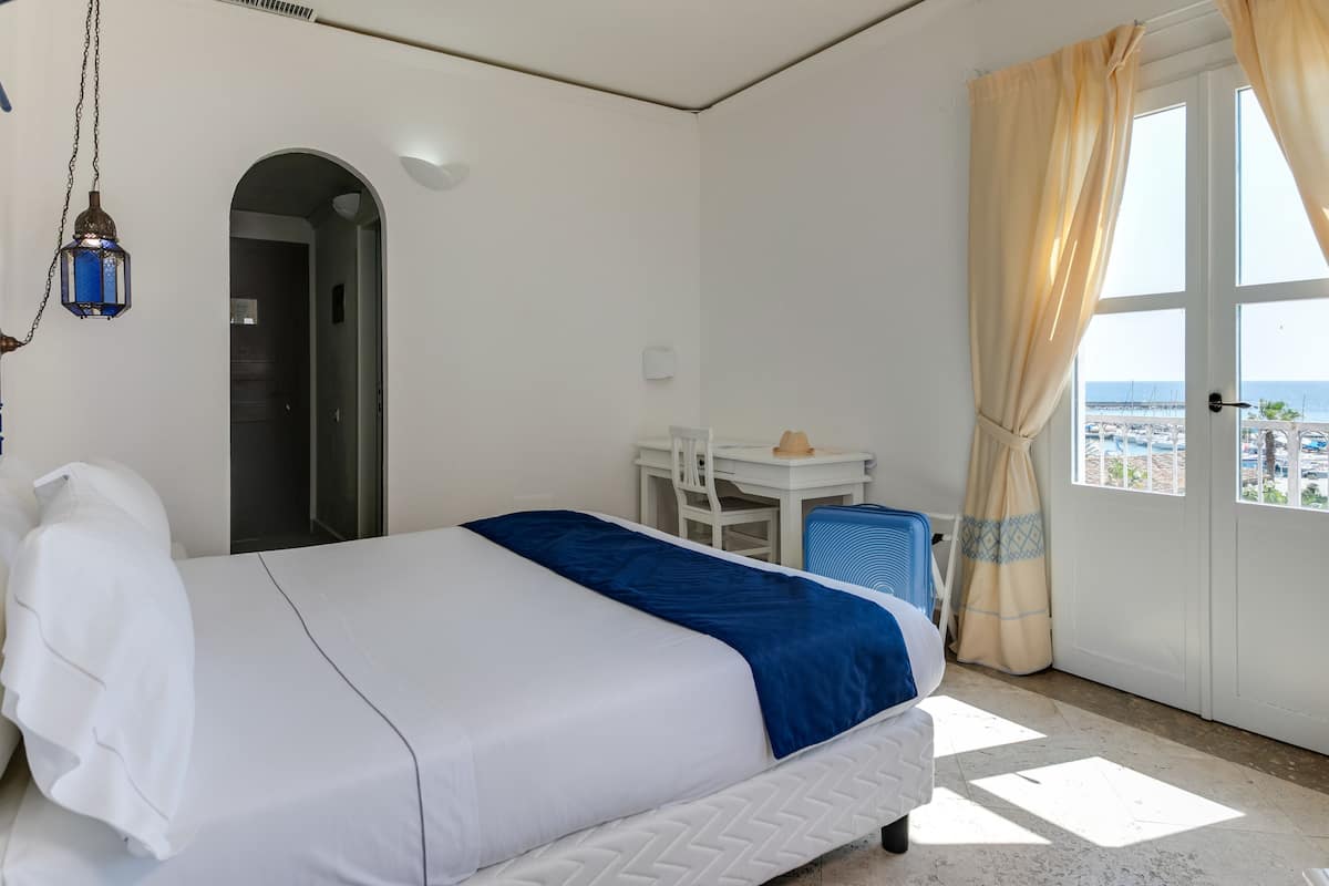 Ein Hotelzimmer mit Bett, Schreibtisch, Stuhl, kleinem Tisch, Pendelleuchte und Meerblick durch ein Fenster.