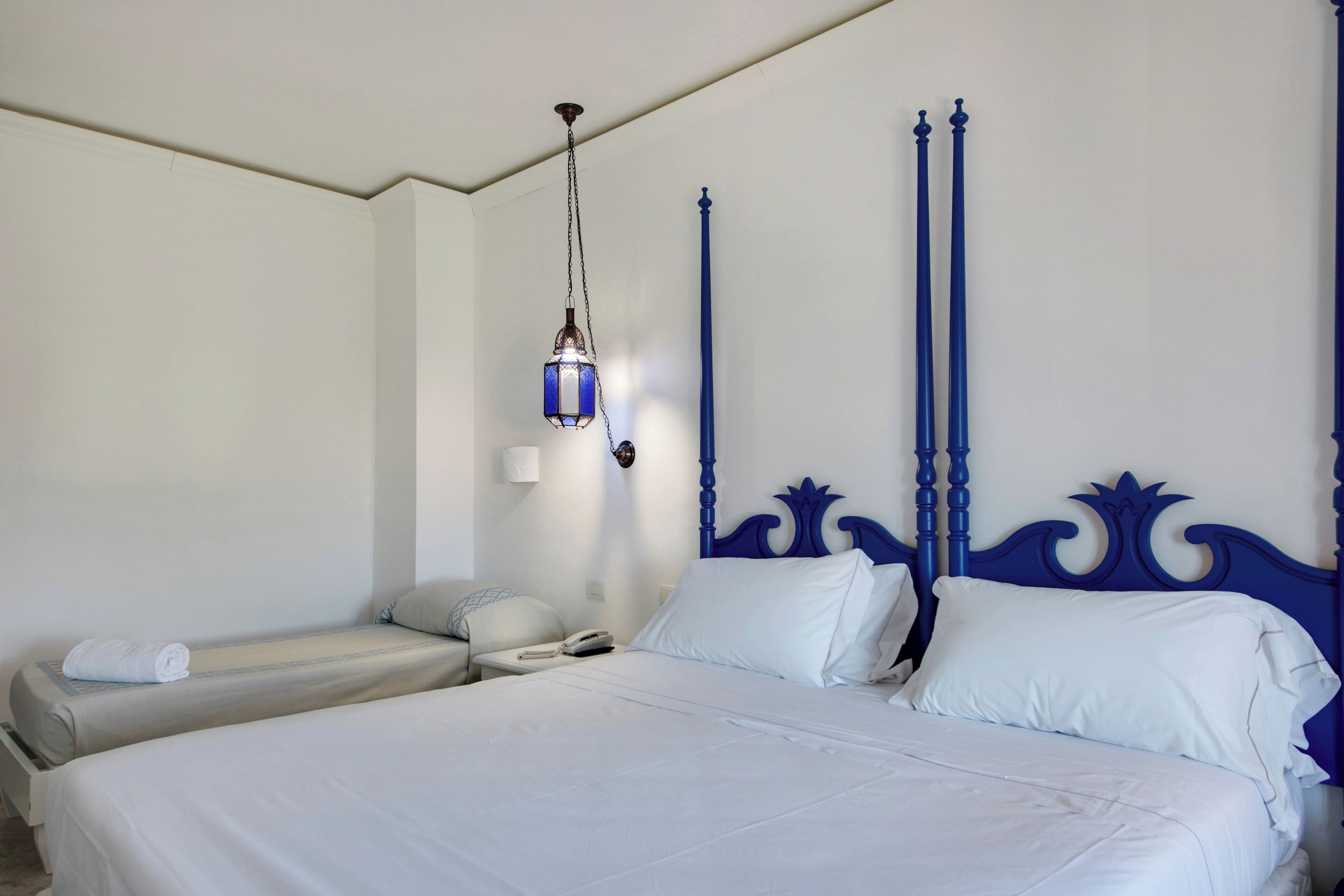 Habitación de hotel con una cama grande, una cama más pequeña, una mesita de noche, una lámpara y un teléfono.