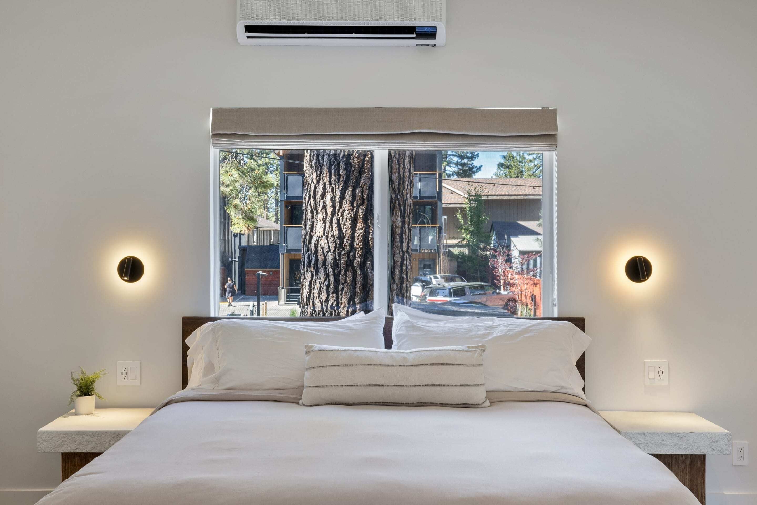 A bedroom with a large window, a bed with white linens, two bedside tables, and wall-mounted lights.