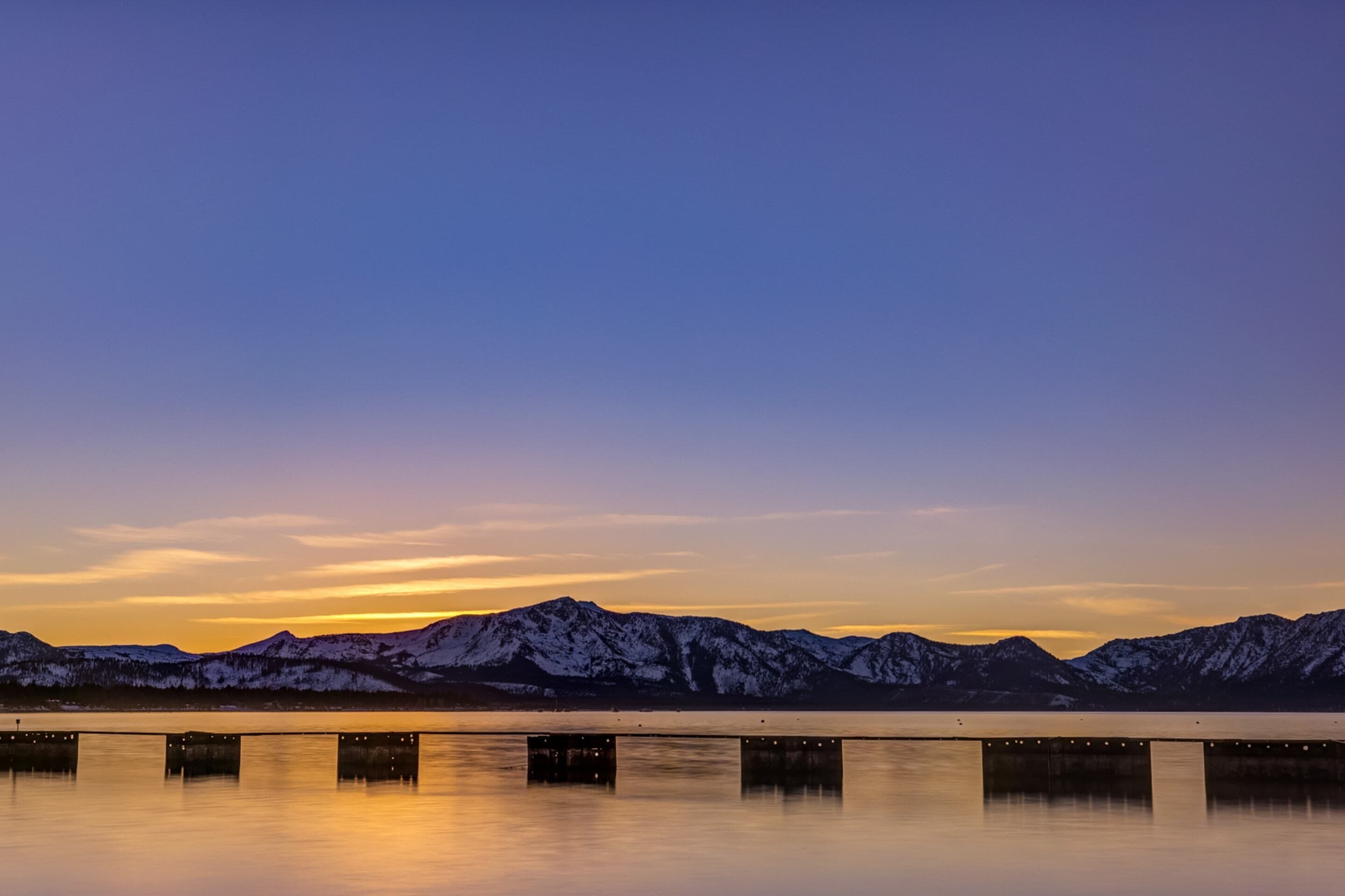 A calm lake with snow-capped mountains in the background and a bridge with lights in the foreground.