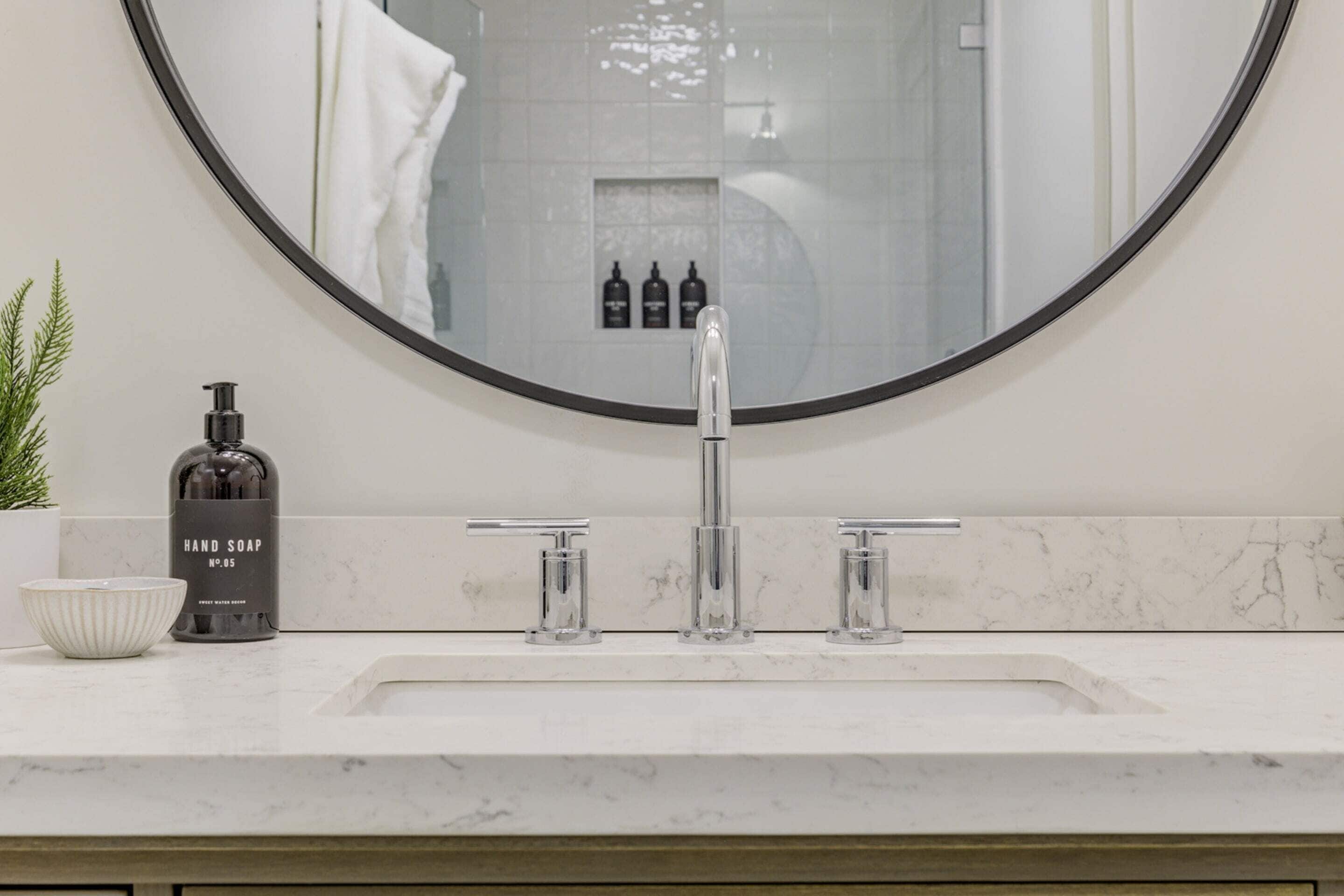 A modern bathroom with a round mirror, a white marble sink, and a hand soap dispenser.