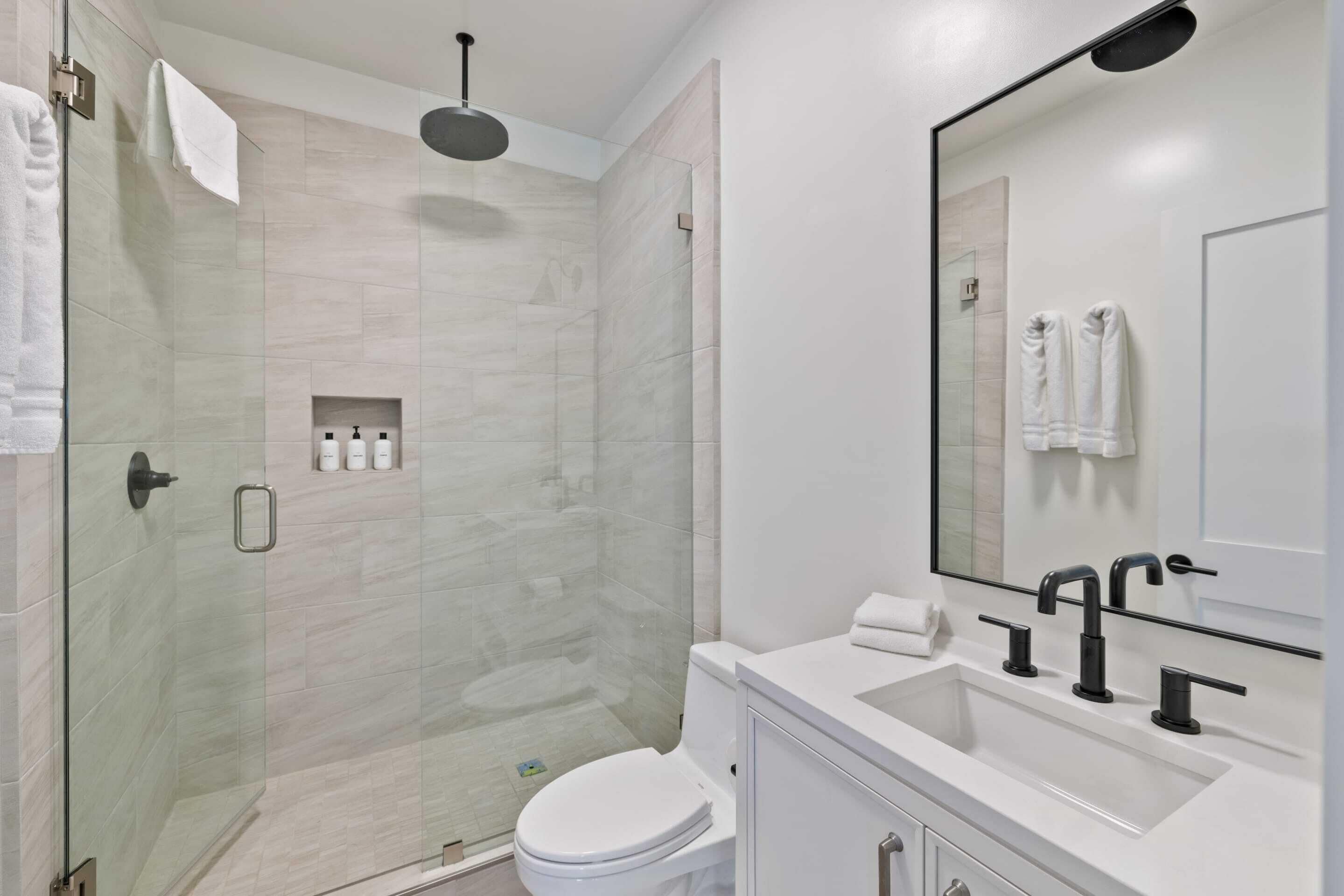 Un baño moderno con ducha de vidrio, inodoro blanco y un espejo grande sobre un lavamanos blanco.
