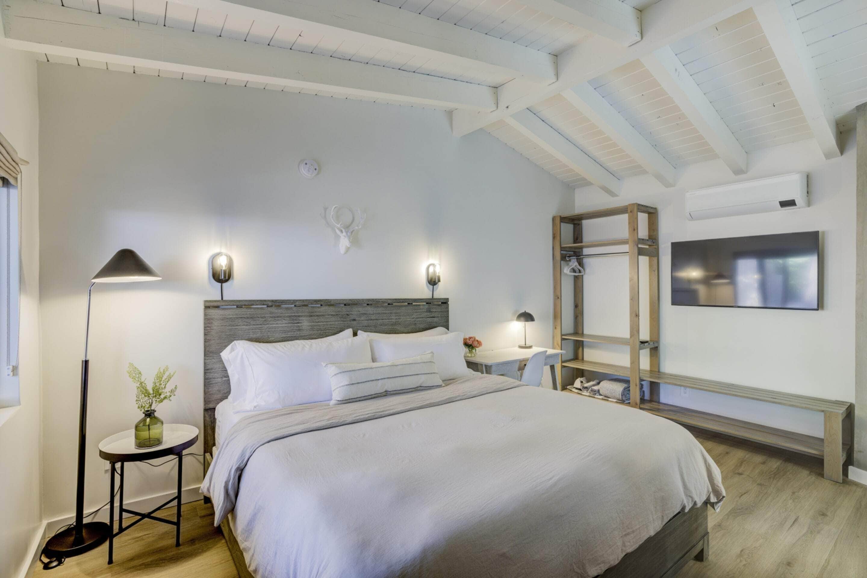 A bedroom with a large bed, a bedside table with a lamp, a small side table with a vase, a wall-mounted TV, and a wooden bench.