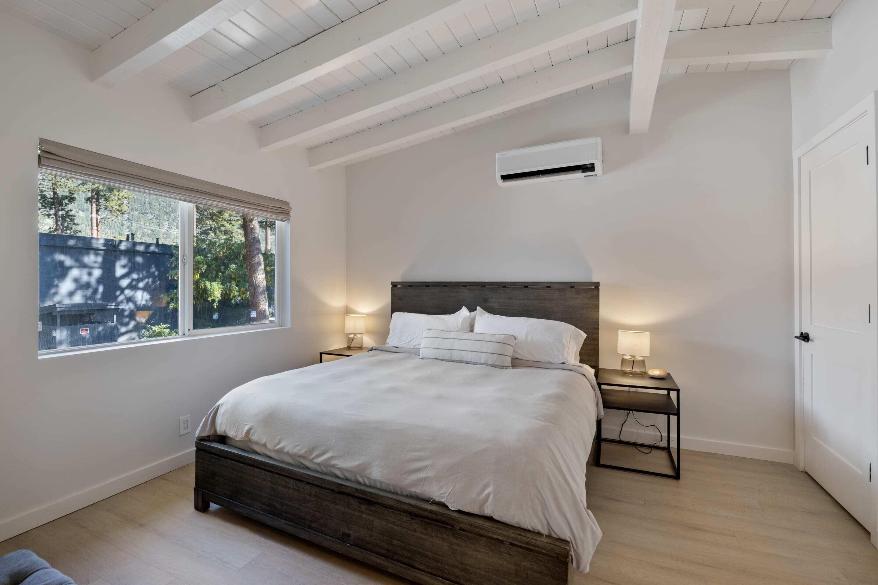 A bedroom with a large bed, two bedside tables with lamps, a window with blinds, and a door.