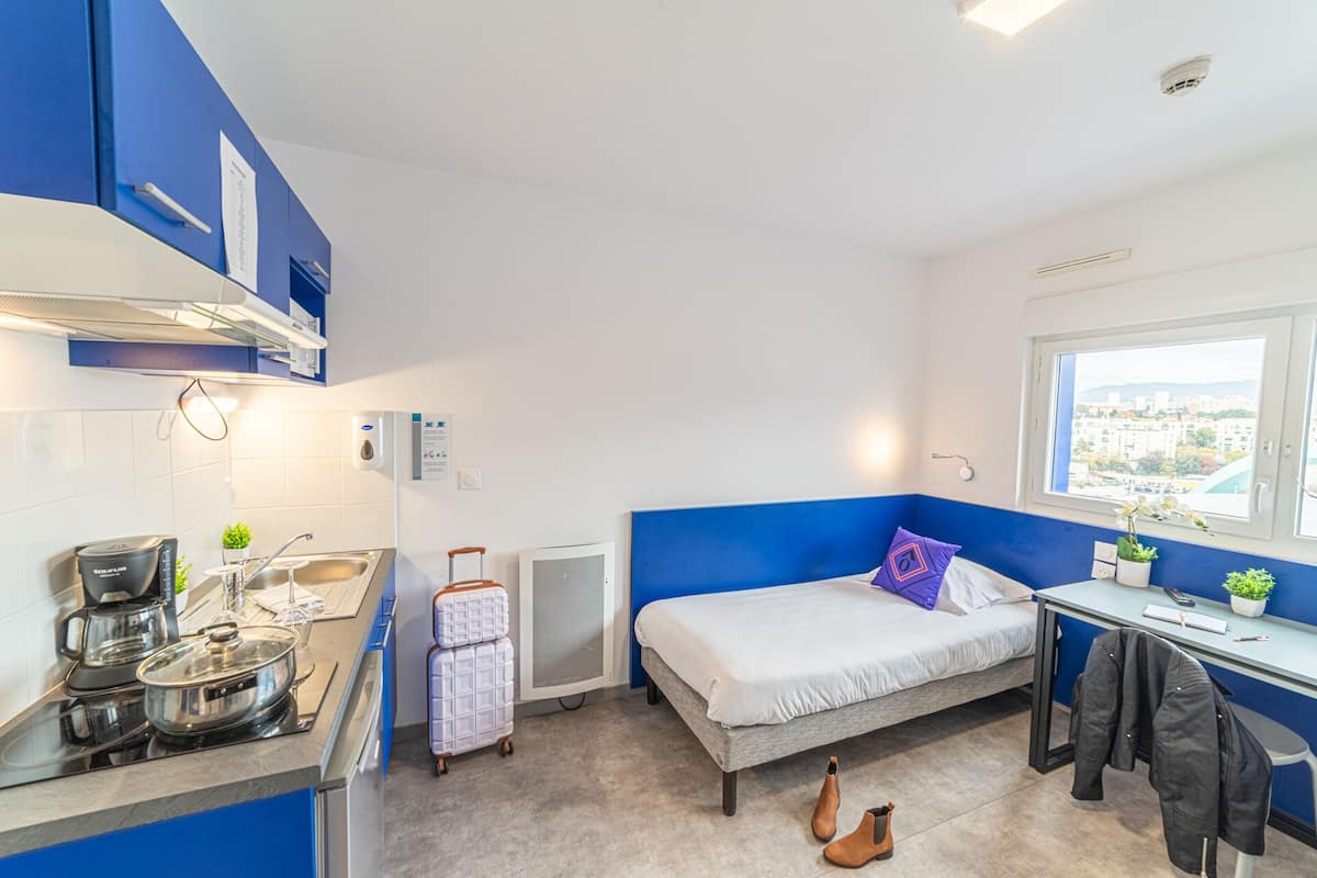 Habitación de hotel compacta con kitchenette, cama con una pared de acento azul, escritorio y ventana con vista a la ciudad.