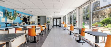 Una cafetería moderna con mesas y sillas altas, una pared de acento azul y amplias ventanas con vistas a zonas verdes.