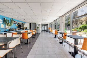 Eine moderne Cafeteria mit hohen Tischen und Stühlen, einer blauen Akzentwand und großen Fenstern mit Blick auf Grünflächen.
