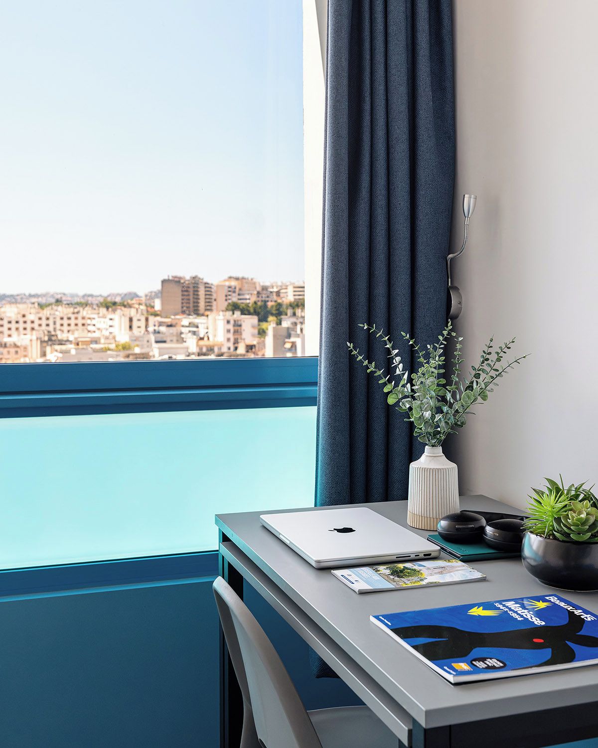A modern room with a view of a cityscape, a desk with a laptop, a plant, and a magazine.