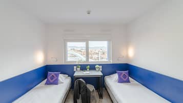 A small, brightly lit room with two beds, a desk, and a window offering a city view.