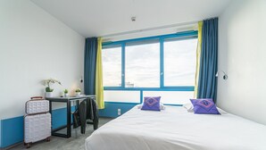 Habitación de hotel con cama, escritorio, silla, maleta y ventana con cortinas.