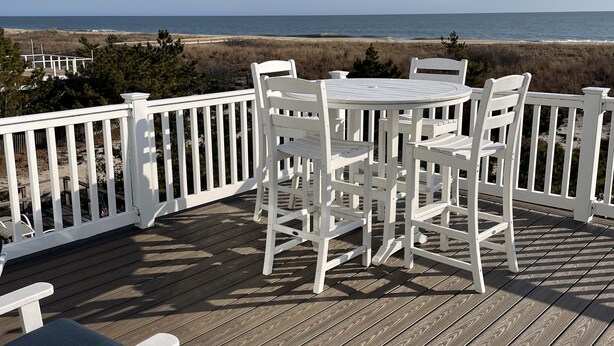 Een houten terras met een witte tafel en stoelen, uitkijkend op een strand.