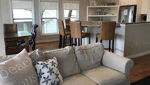 Een moderne keuken met witte kasten, een granieten aanrechtblad en een roestvrijstalen koelkast. Er is een zithoek met een houten tafel en stoelen, en een woonkamer met een beige bank en een decoratief kussen.