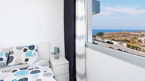 Une chambre d’hôtel moderne équipée d’un lit, d’une table de chevet, d’une lampe et d’une grande fenêtre donnant sur la mer et les bâtiments environnants.