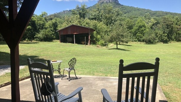Een overdekt terras met twee houten stoelen en een kleine tafel, met uitzicht op een gazon met een rood gebouw en een berg op de achtergrond.
