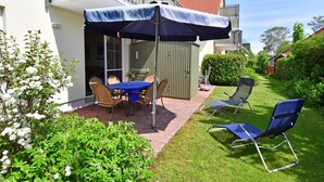 Un patio al aire libre con una mesa y sillas bajo un paraguas azul, rodeado de follaje y un edificio con tejado de tejas rojas.