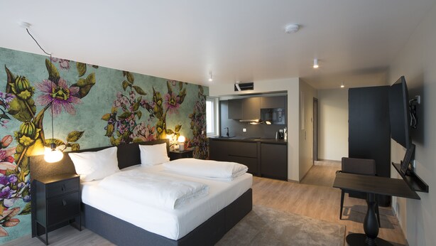 Une chambre moderne avec un grand lit, des tables de chevet, une télévision et un coin cuisine à l’arrière-plan.