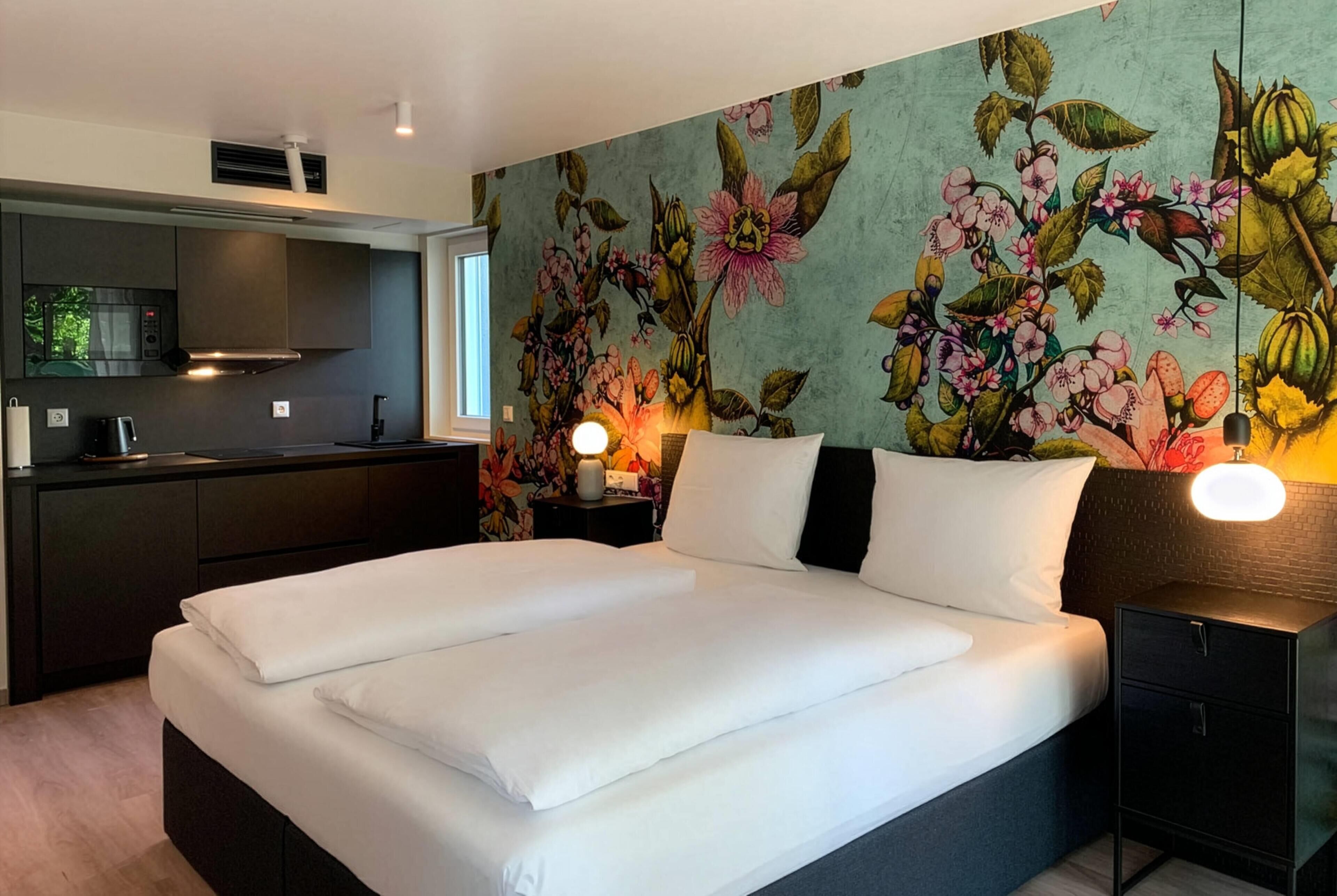 Un dormitorio moderno con una cama grande, una mesita de noche con lámpara, un escritorio empotrado con microondas y una pared con un mural floral.