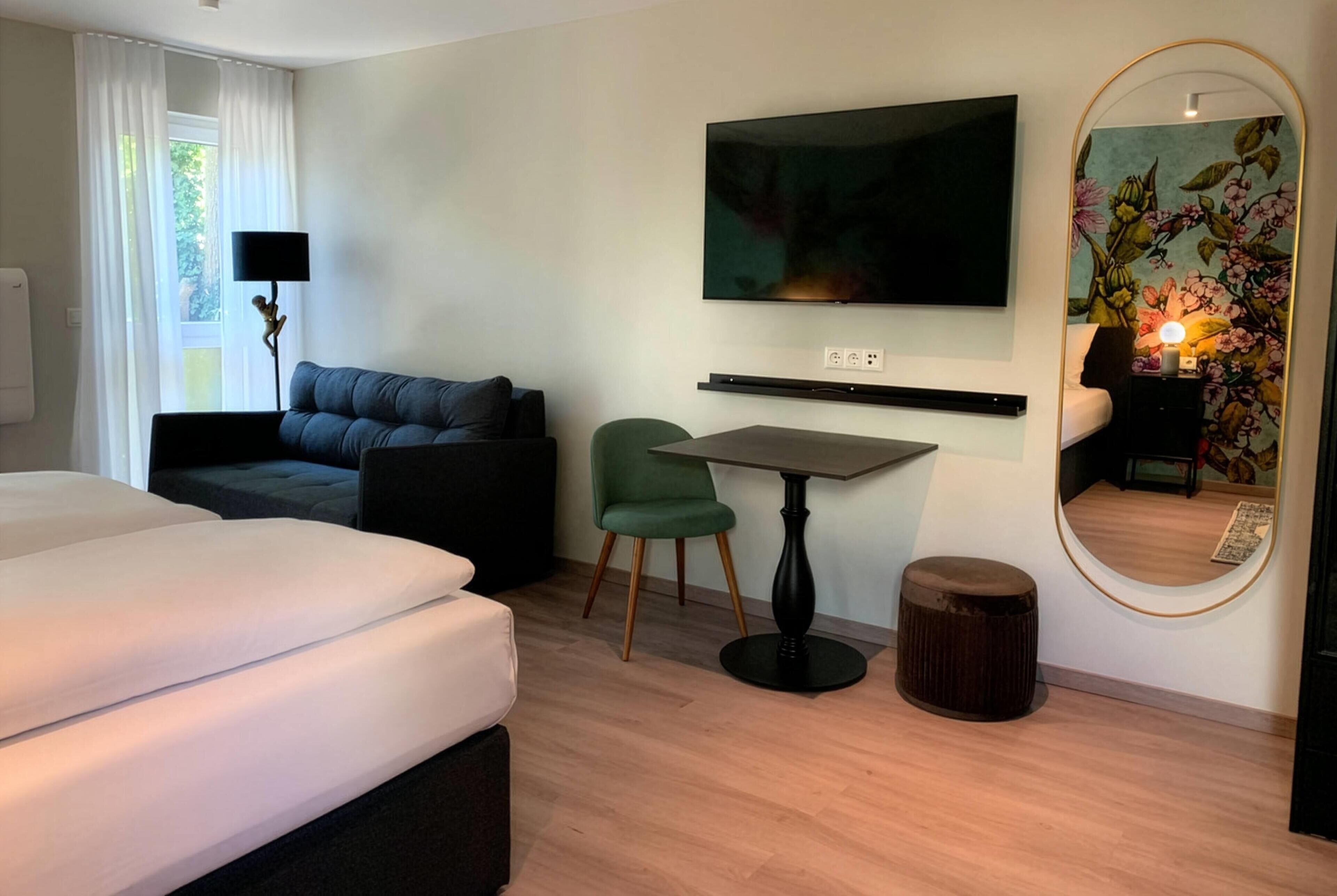 Habitación de hotel con cama, sofá, escritorio, silla, televisor, espejo y piso de madera.