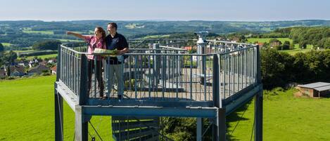 A metal observation tower with a staircase and a railing, overlooking a green landscape with houses.