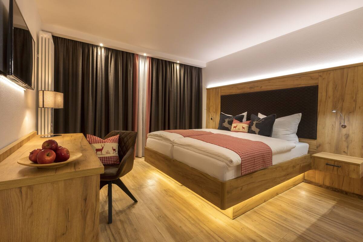Quarto de hotel moderno com cama de madeira, escrivaninha com maçãs, cadeira e uma janela grande com cortinas.