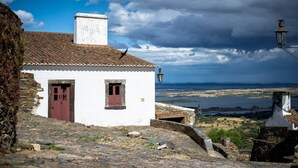 Um edifício branco com porta e janelas vermelhas, com vista para um vale com rio e montanhas ao longe.