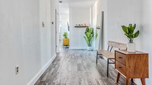 Un pasillo estrecho con suelo de madera, un banco, una mesita de madera con una planta y un aparador amarillo al fondo.