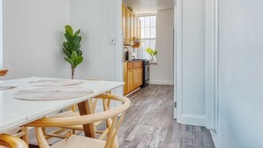 Un comedor con una mesa blanca y sillas de madera, una cocina al fondo y una planta sobre la mesa.