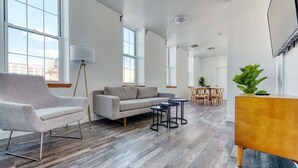 Un salón amplio con un sofá gris, un sillón gris y una pequeña mesa de centro. Se observa un aparador de madera con una planta encima.