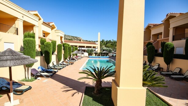 A pool area with lounge chairs and umbrellas, surrounded by neatly trimmed hedges and palm trees. - LUXURY APARTMENT - T1 pool view - PRAIA DA LUZ (Luz)