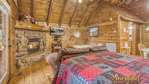 Une cabane avec une cheminée en pierre, des murs en bois, une courtepointe, un ventilateur de plafond et un tableau accroché au mur.