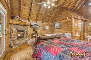 A cabin with a stone fireplace, wooden walls, a quilted bedspread, a ceiling fan, and a mounted picture. - Cozy one bedroom studio style cabin (Sevierville)