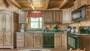 A wooden cabin kitchen with a large window, wooden cabinets, a microwave, and a dishwasher. - Located in a peaceful setting, convenient to both Gatlinburg and Pigeon Forge! (Sevierville)