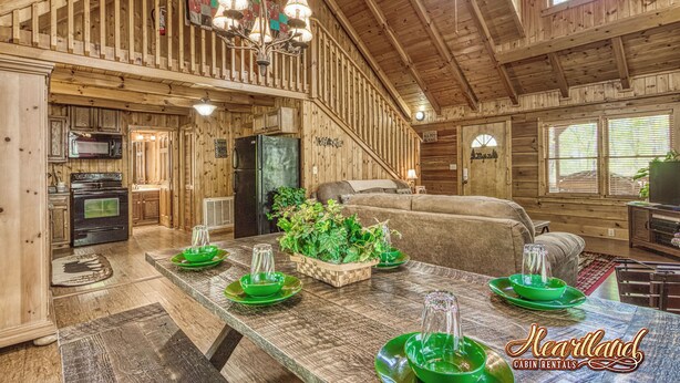 A cabin interior with a wooden staircase, a dining area with a large table, and a kitchen with modern appliances. - Located in a peaceful setting, convenient to both Gatlinburg and Pigeon Forge! (Sevierville)